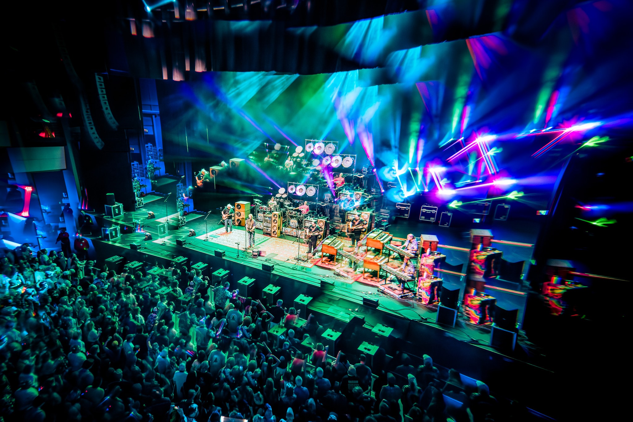 Overhead wide shot of full band on stage with colorful green and blue light beams and crowd